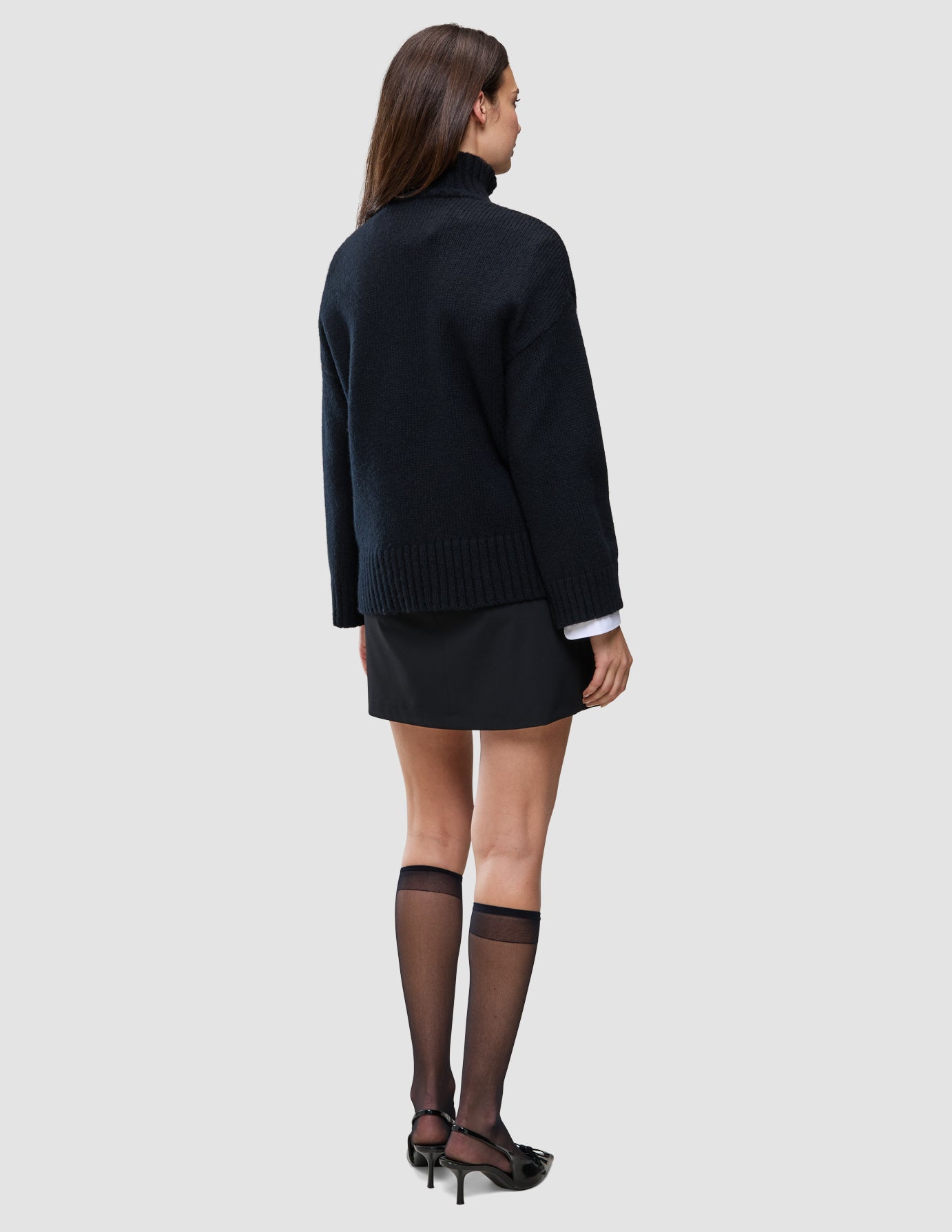 En person sett bakfra, kledd i en svart, tykk og myk ullstrikket turtleneck for kvinner, svart skjørt og gjennomsiktige knestrømper.
